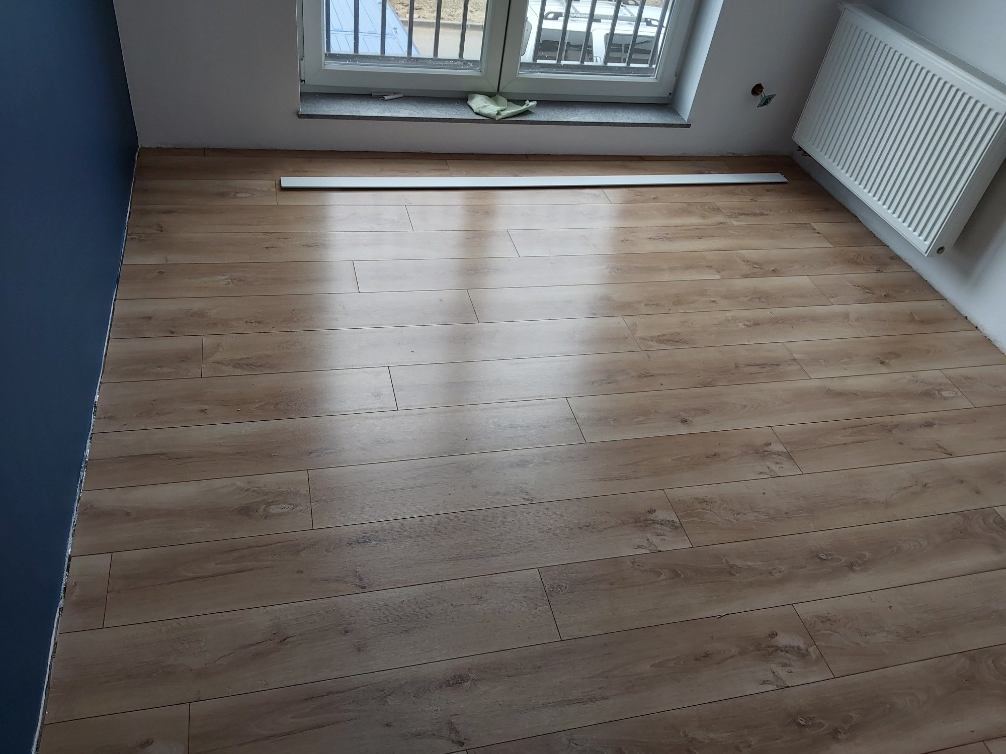 Ułożone panele podłogowe o wzorze drewna w pokoju z niebieską ścianą, białą listwą przypodłogową i grzejnikiem. Widoczne okno z parapetem i fragmentem ulicy za nim.