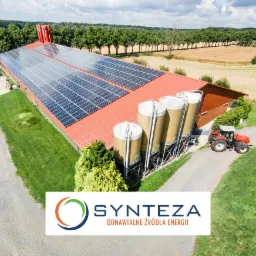 Instalacja fotowoltaiczna na dachu budynku gospodarczego z silosami i czerwonym traktorem, widok z lotu ptaka, logo firmy w dolnej części kadru.