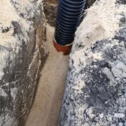 Rura karbowana w wykopie wypełnionym piaskiem, widoczne ślady po pracy koparki na ścianach wykopu.