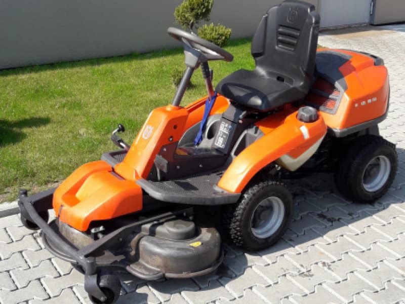 Pomarańczowy traktorek ogrodowy Husqvarna Rider na kostce brukowej, z fragmentem trawnika w tle.