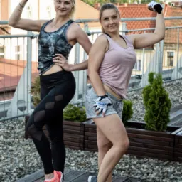 Dwie uśmiechnięte kobiety w sportowych strojach prezentują swoje bicepsy na tle miejskiej zabudowy i zieleni, stojąc na drewnianym tarasie.