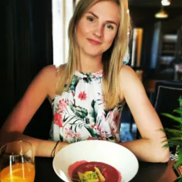 Uśmiechnięta blondynka w kwiecistej sukience siedzi przy stole z talerzem czerwonego barszczu z tortellini i szklanką soku pomarańczowego.