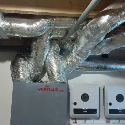 Szare urządzenie Ventilus 390 z czerwonym napisem, podłączone do srebrnych, elastycznych rur wentylacyjnych pod sufitem z drewnianymi belkami, obok dwa białe falowniki Fronius SYMO na ścianie.