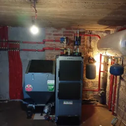 Kotłownia w Łobodnie z nowoczesnym piecem na paliwo stałe, zbiornikiem buforowym i rozdzielaczem instalacji grzewczej z czerwonymi rurami.