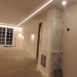 Damiano - Wnętrze w trakcie prac wykończeniowych: ściany po nałożeniu gładzi, zabezpieczony kominek folią, widoczny osuszacz powietrza Master.