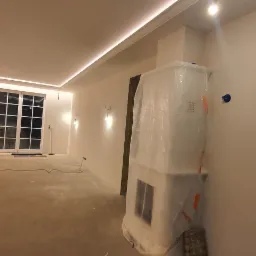 Wnętrze w trakcie prac wykończeniowych: ściany po nałożeniu gładzi, zabezpieczony kominek folią, widoczny osuszacz powietrza Master.