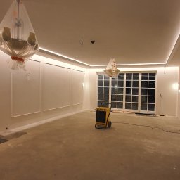 Damiano - Wnętrze w trakcie remontu z osuszaczem powietrza, ściany z dekoracyjnymi listwami, zakryte folią, widoczne przygotowania do malowania, oświetlenie LED w suficie.