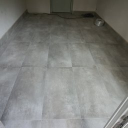 Damiano - Nowo ułożone, szare płytki podłogowe w pustym pomieszczeniu, widoczne z perspektywy otwartych drzwi, z niebieską rurą i butelką wody przy ścianie.