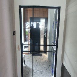 Damiano - Nowoczesne, czarne, przeszklone drzwi wewnętrzne z podziałami, widoczne w trakcie montażu, zabezpieczone folią ochronną, z widocznymi śladami prac wykończeniowych na podłodze i ścianach.