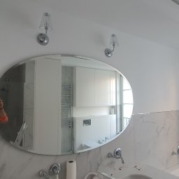 Damiano - Wykończona łazienka z owalnym lustrem, marmurowymi płytkami na ścianach, chromowaną armaturą i oświetleniem ściennym; w lustrze odbicie osoby robiącej zdjęcie.