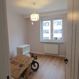 Damiano - Pomalowany na jasny róż pokój z drewnianą podłogą, białym regałem i grzejnikiem pod oknem, częściowo zagracony kartonami i rowerkiem dziecięcym.