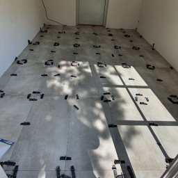 Damiano - Szare płytki podłogowe w trakcie układania, z systemem poziomowania, na podłodze widoczne cienie i fragmenty drzwi.