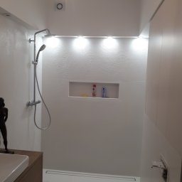 Damiano - Nowoczesna kabina prysznicowa z deszczownicą i odpływem liniowym, białe płytki z teksturą imitującą fale, wnęka z kosmetykami, oświetlenie punktowe i drewniana szafka z umywalką.