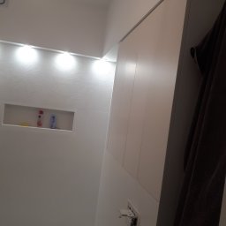 Damiano - Nowoczesna łazienka z oświetleniem LED, dekoracyjną strukturą na ścianie i wnęką z kosmetykami, widoczne gniazdko elektryczne bez obudowy i wiszący brązowy ręcznik.