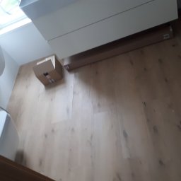 Damiano - Podłoga z jasnych paneli, widoczne kartony i paczka paneli pod szafką, okno dachowe.