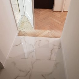 Damiano - Połączenie białych płytek imitujących marmur z drewnianą podłogą ułożoną w jodełkę, widok na otwarte białe drzwi z ciemnym pomieszczeniem w tle.