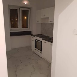 Damiano - Wykończona kuchnia w odcieniach bieli z marmurową podłogą i blatem, widoczna przez wejście do pomieszczenia, oświetlona lampą wiszącą.