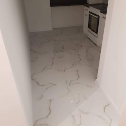 Damiano - Wykończona kuchnia z białymi płytkami podłogowymi imitującymi marmur i widoczną częścią zabudowy kuchennej z białą kuchenką.