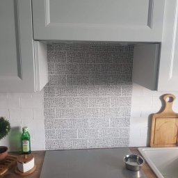 Damiano - Kuchnia z szarymi szafkami, białymi płytkami i panelem z motywem typograficznym nad blatem, widoczna deska do krojenia i akcesoria kuchenne.