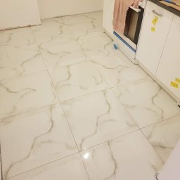 Damiano - Błyszcząca podłoga wyłożona białymi płytkami imitującymi marmur, świeżo po ułożeniu, widoczne fragmenty ścian i szafek kuchennych z zabezpieczeniami.