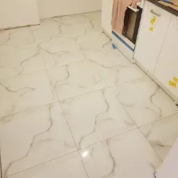 Błyszcząca podłoga wyłożona białymi płytkami imitującymi marmur, świeżo po ułożeniu, widoczne fragmenty ścian i szafek kuchennych z zabezpieczeniami.