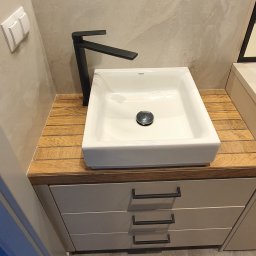 Damiano - Nowoczesna, kwadratowa umywalka Grohe z czarną armaturą na drewnianym blacie z szufladami w łazience, widoczne odbicie osoby robiącej zdjęcie w lustrze.