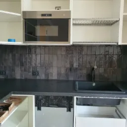 Częściowo wykończona kuchnia z czarnym blatem, ciemnymi płytkami na ścianie, zamontowanym piekarnikiem i szafkami bez frontów, ukazująca etap prac.