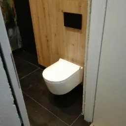 Nowoczesna, podwieszana toaleta w łazience z drewnianą zabudową i czarnymi płytkami podłogowymi, widoczna z perspektywy wejścia, z niedokończonym wykończeniem framugi drzwi.