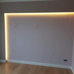 Damiano - Wnęka w ścianie pomalowanej na jasny fiolet z podświetleniem LED w ciepłym odcieniu, widoczne gniazdka elektryczne i drewniana podłoga.