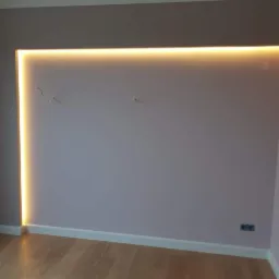 Wnęka w ścianie pomalowanej na jasny fiolet z podświetleniem LED w ciepłym odcieniu, widoczne gniazdka elektryczne i drewniana podłoga.