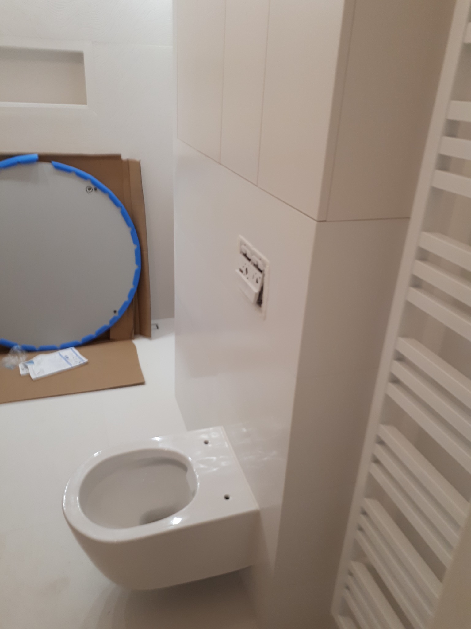Wisząca miska WC zamontowana w białej łazience, widoczny karton z okrągłym lustrem z niebieską obwódką i grzejnik drabinkowy.