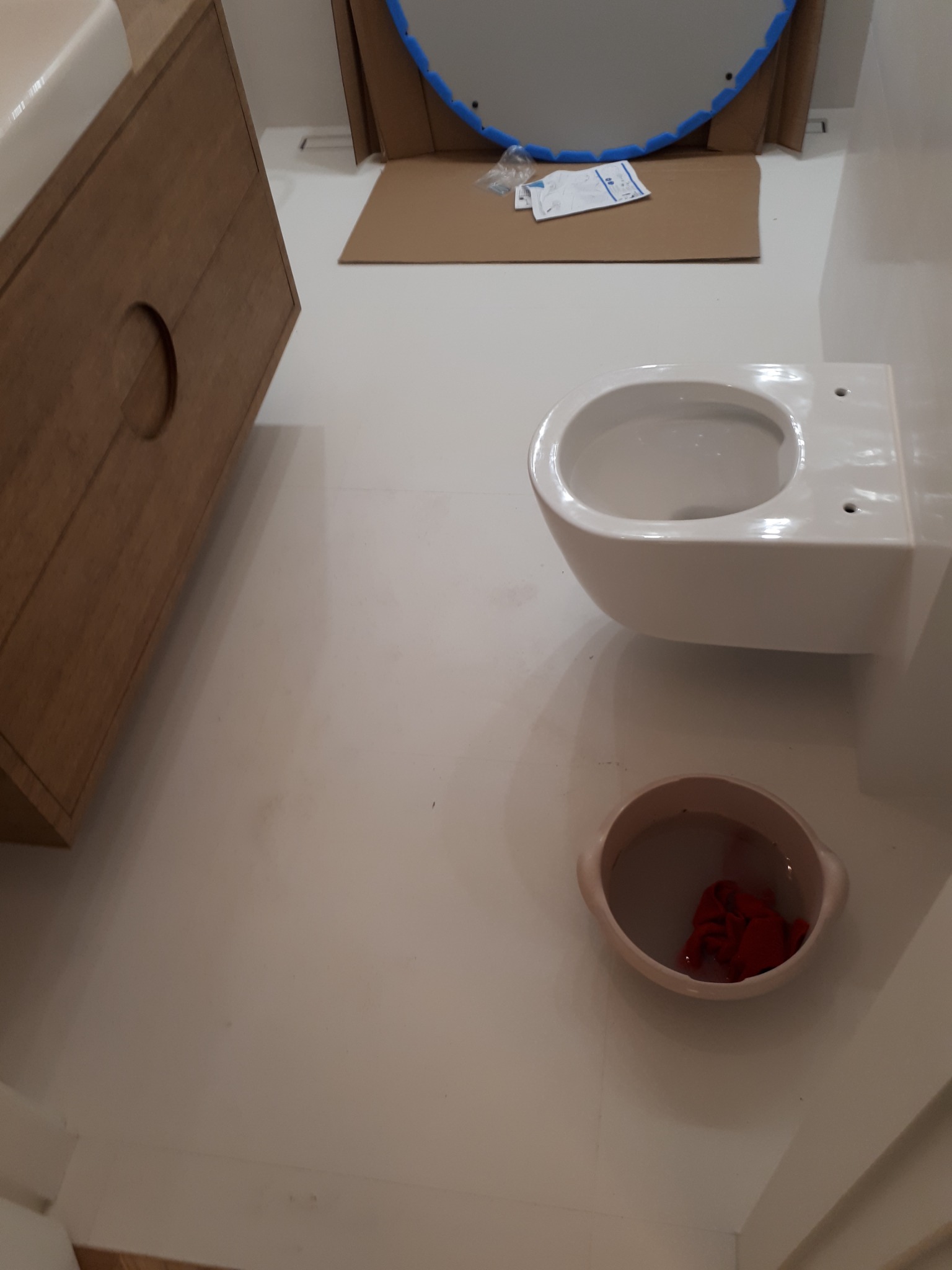 Biała, podwieszana miska WC przed montażem, obok umywalka z szafką z jasnego drewna, na podłodze karton z lustrem w niebieskiej ramie i miska z czerwoną szmatką.