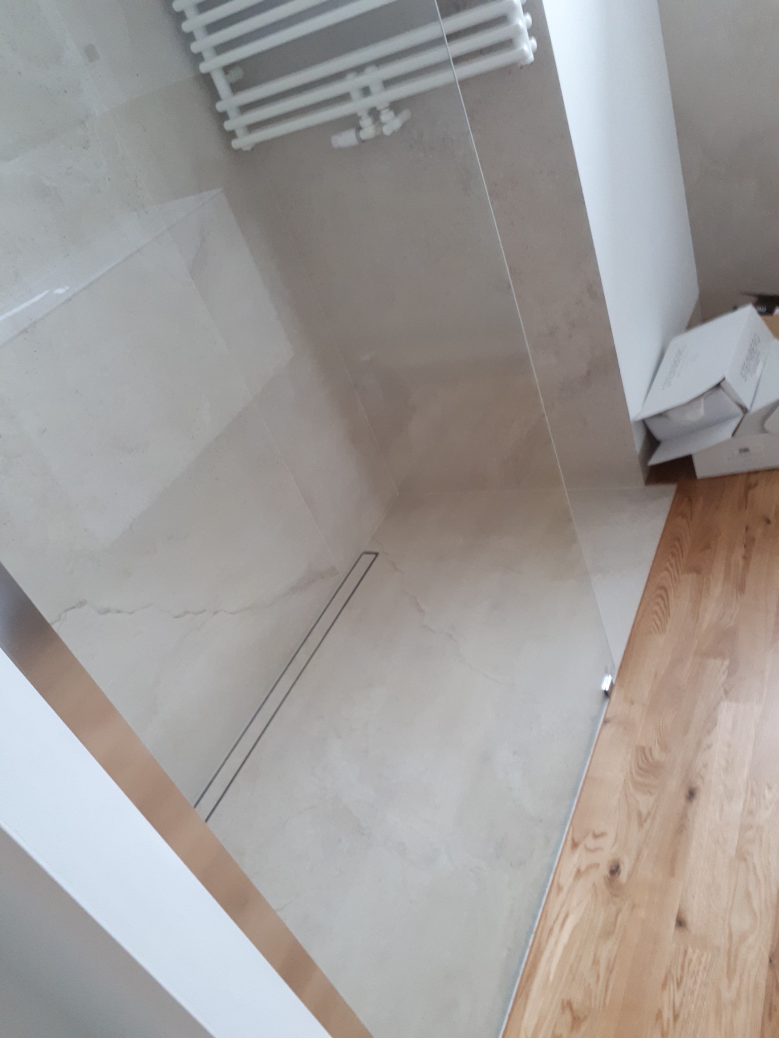 Nowoczesna kabina prysznicowa z odpływem liniowym, wykończona płytkami imitującymi marmur i drewnianą podłogą, widoczny grzejnik drabinkowy i kartony w tle.