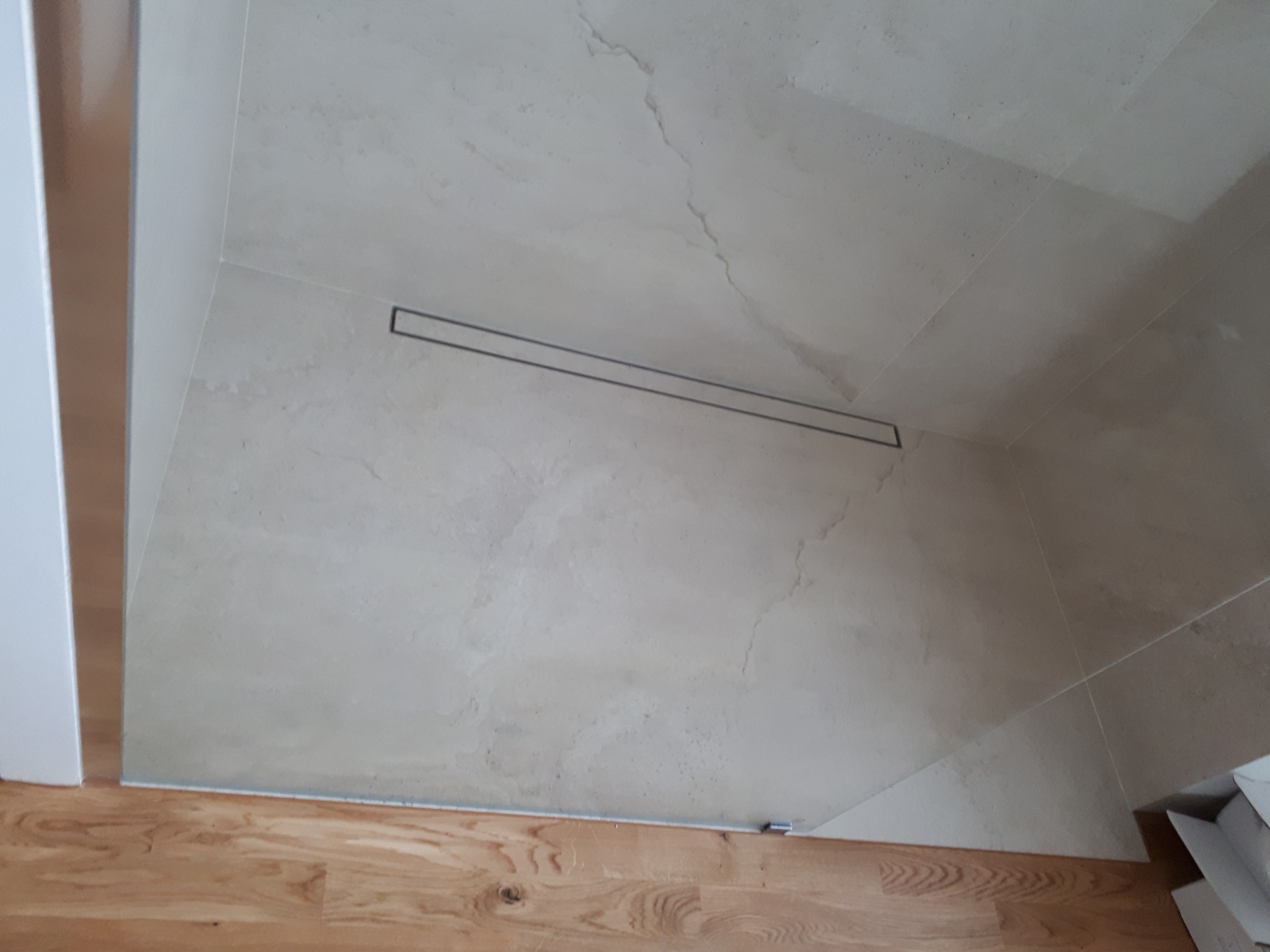 Wykończona strefa prysznicowa z beżowymi płytkami imitującymi marmur i odpływem liniowym, widoczna część podłogi z drewnianych paneli.