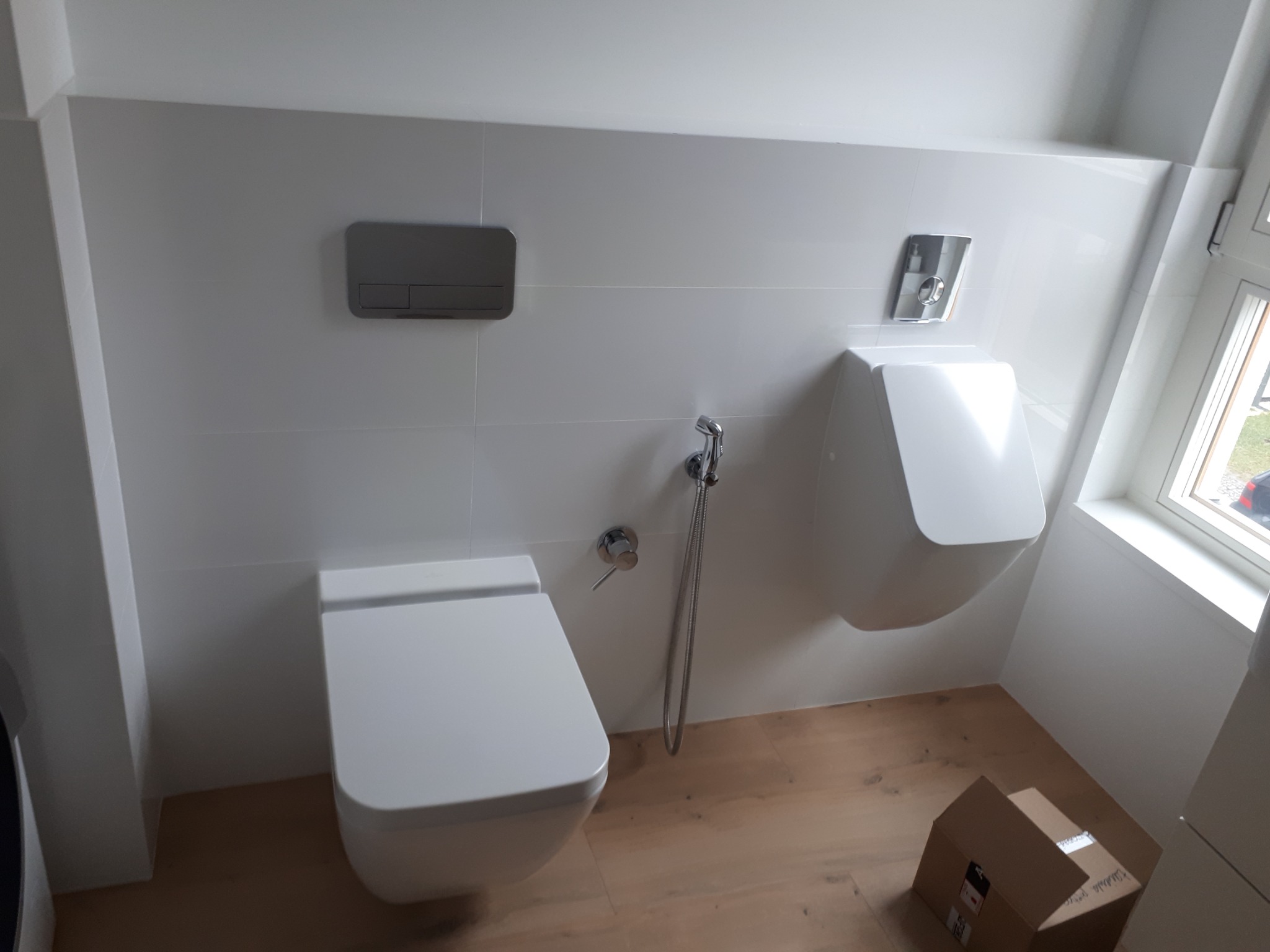 Nowoczesna, minimalistyczna łazienka z białymi płytkami, wiszącą toaletą i pisuar, drewnopodobna podłoga i kartonowe pudełka w rogu.