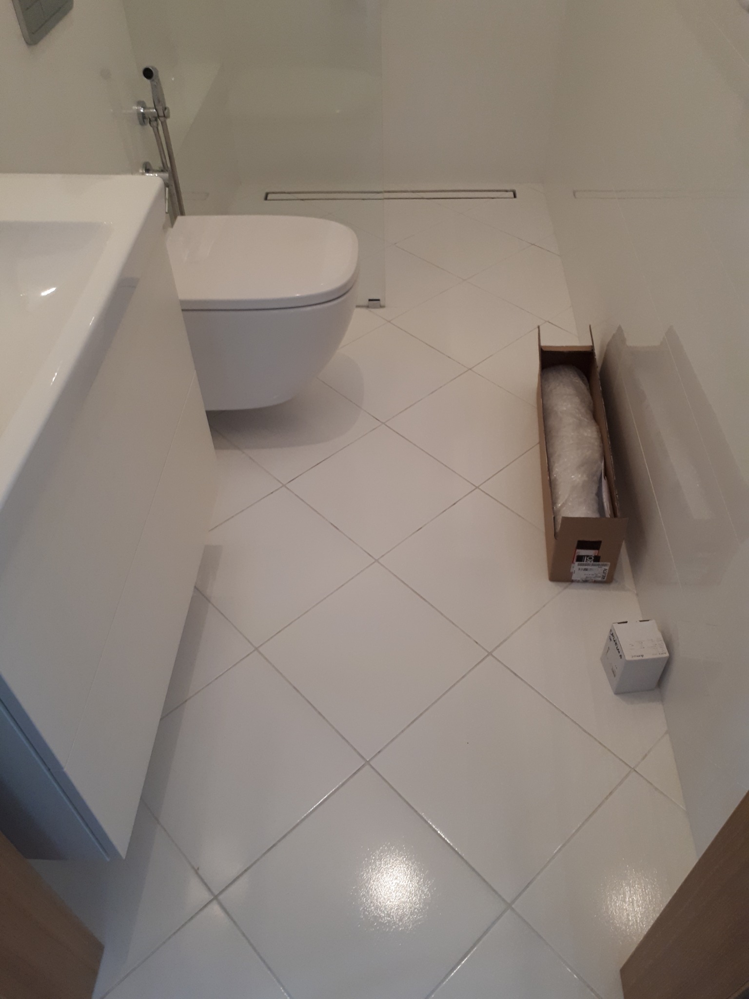 Nowoczesna łazienka z białą armaturą, płytkami podłogowymi ułożonymi w karo oraz kartonem z zapakowanym elementem wyposażenia.