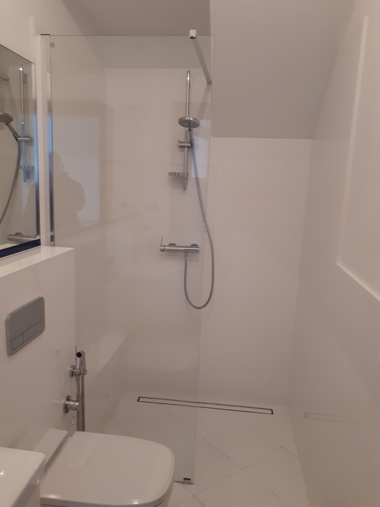 Nowoczesna, minimalistyczna łazienka z kabiną prysznicową typu walk-in, odpływem liniowym w podłodze i toaletą z bidetką, utrzymana w bieli.