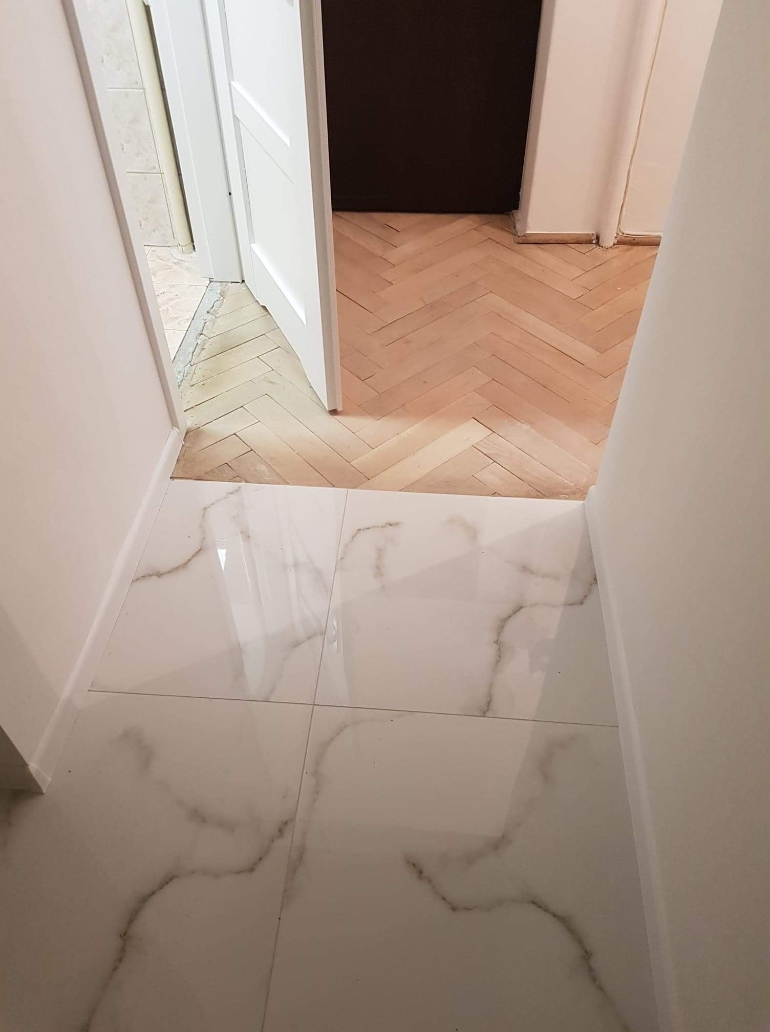 Połączenie białych płytek imitujących marmur z drewnianą podłogą ułożoną w jodełkę, widok na otwarte białe drzwi z ciemnym pomieszczeniem w tle.