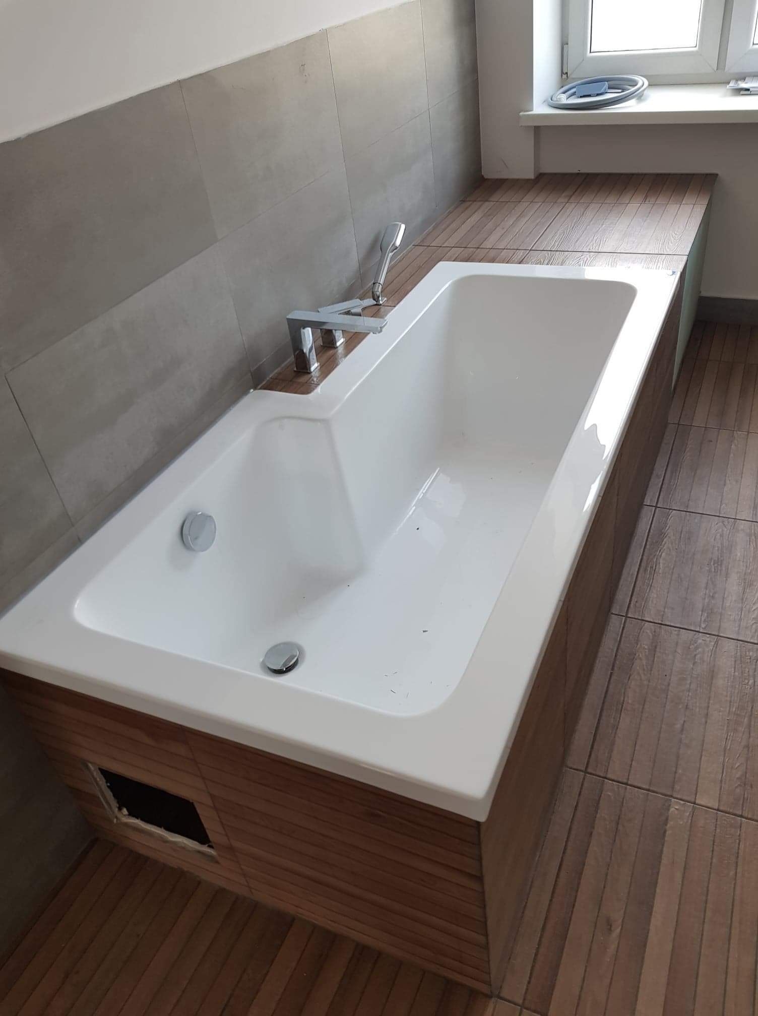 Nowoczesna wanna obudowana drewnopodobnymi panelami, z chromowaną armaturą i szarą ścianą w tle, widoczny otwór rewizyjny w obudowie wanny.