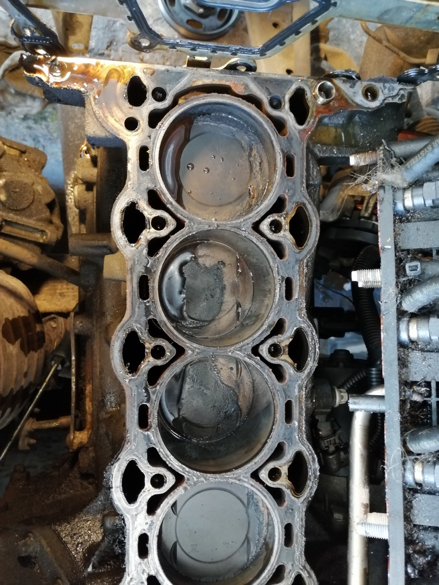 Zdjęcie bloku silnika samochodowego z widocznymi cylindrami, z śladami oleju i płynów, prawdopodobnie w trakcie demontażu lub naprawy.