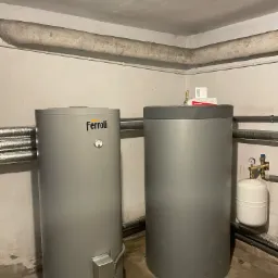 Dwa szare zasobniki ciepłej wody, jeden marki Ferroli, w piwnicy z rurami grzewczymi i betonowym stropem.