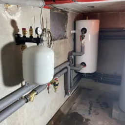 Biały zbiornik wyrównawczy ciśnienia z manometrem, zamontowany na ścianie, podłączony do rur w szarej izolacji, obok biały bojler w surowym pomieszczeniu technicznym.