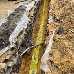 Wykop w ziemi zabezpieczony żółtą taśmą ostrzegawczą z napisem 'GAZ', widoczne rury i kostka brukowa zabezpieczona folią, w tle ceglany budynek.