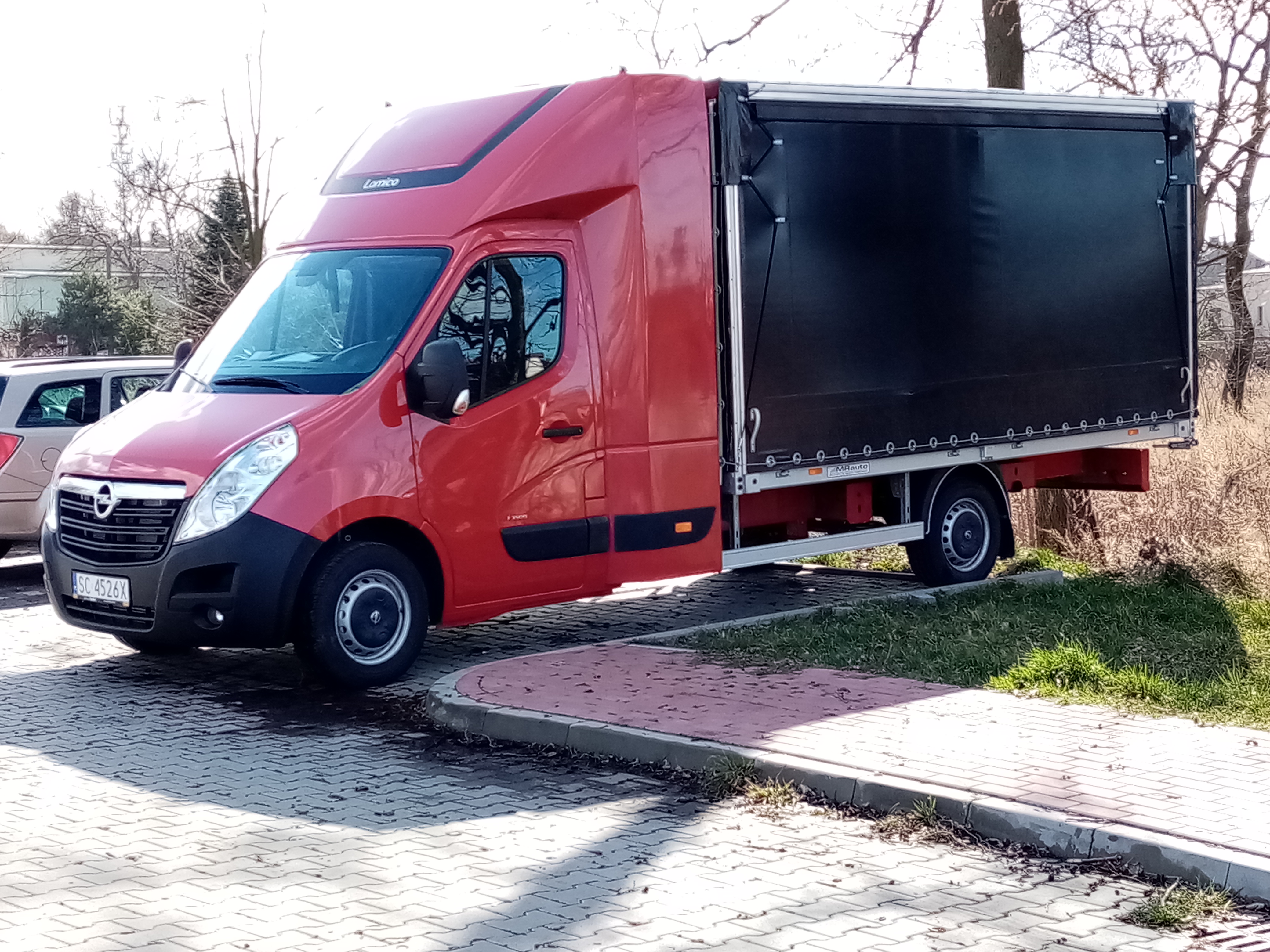 Czerwony bus dostawczy z czarną plandeką na parkingu z kostki brukowej, widoczna tablica rejestracyjna i logo Lamico.