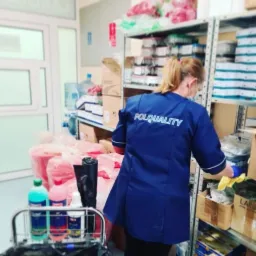 Kobieta w niebieskim uniformie z napisem POLQUALITY, w żółtych rękawiczkach, porządkuje półki w magazynie, wózek z detergentami obok, widoczne butelki z płynami do czyszczenia.