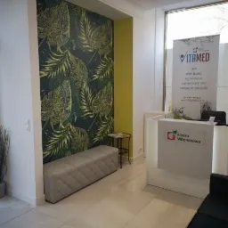 Recepcja w gabinecie lekarskim z tapetą w motywy roślinne, białą ladą z logo 'Klinika Witaminowa' i szarym siedziskiem.