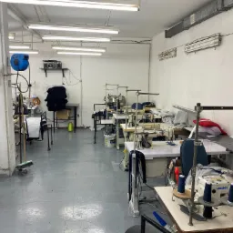 Przestronna szwalnia z kilkoma stanowiskami do szycia, maszyny JUKI, stosy materiałów i gotowych ubrań, widoczny gaśnica i radio na półce