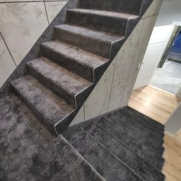 DANIEL ADAMSKI PODŁOGI - Schody wyłożone szarą wykładziną dywanową z ciemnym obramowaniem, widok z góry. W tle jasna ściana z efektem betonu i fragment podłogi z paneli.