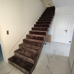 DANIEL ADAMSKI PODŁOGI - Schody wyłożone brązową wykładziną dywanową na białych marmurowych płytkach. Nowoczesne wnętrze z minimalistyczną balustradą i białymi ścianami.