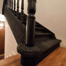 DANIEL ADAMSKI PODŁOGI - Schody wyłożone ciemną, teksturowaną wykładziną, z czarnymi drewnianymi balustradami. Widoczny fragment drewnianej podłogi na dole schodów.